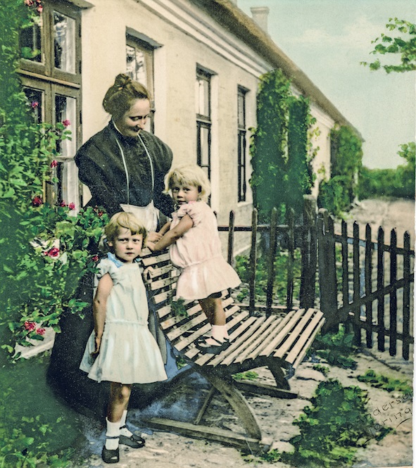 Lærerinde Karen Andersen med førstelærer Johannes Nørgaards to ældste børn foran den gamle skole i Øster Hassing i 1911. Håndkoloreret foto taget af Hjørring-fotografen Helga Andersen. B13826.